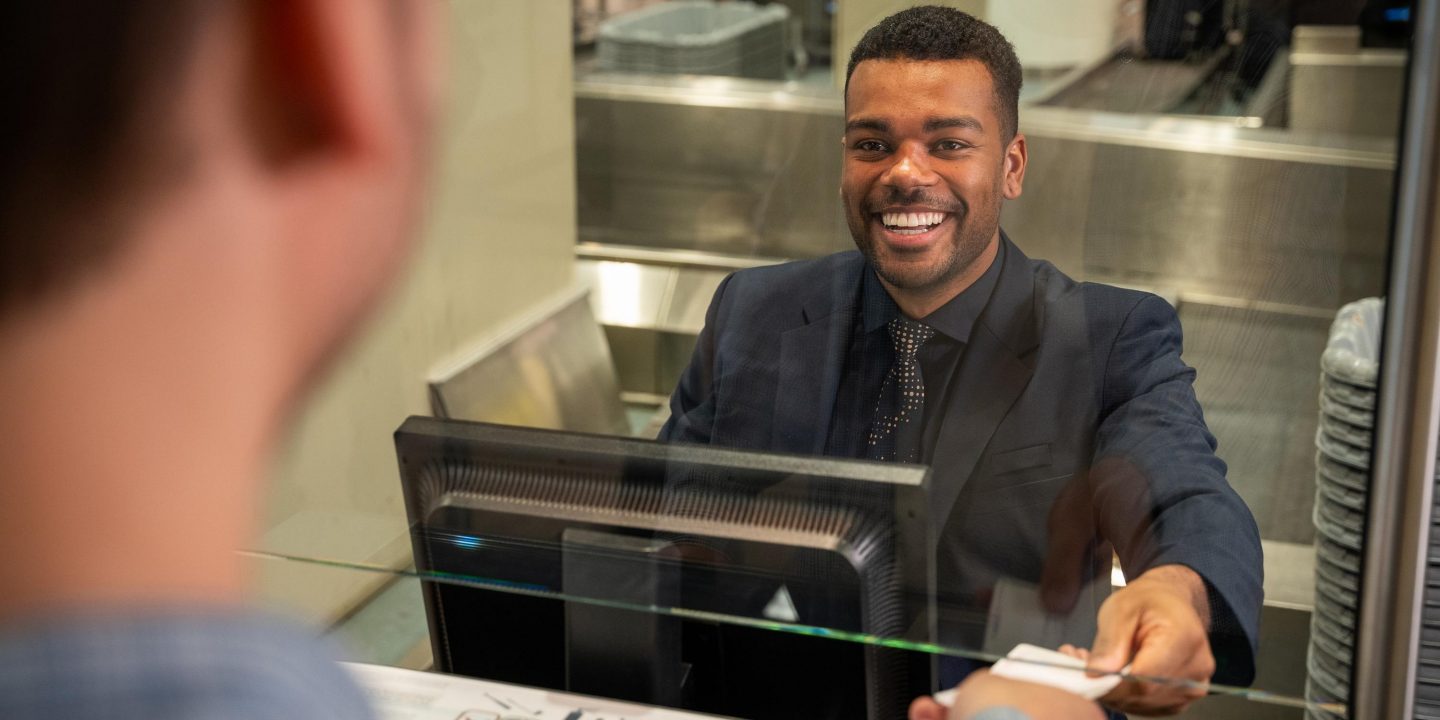 A smiling employee in a suit hands a document to a customer over a counter with a glass partition.