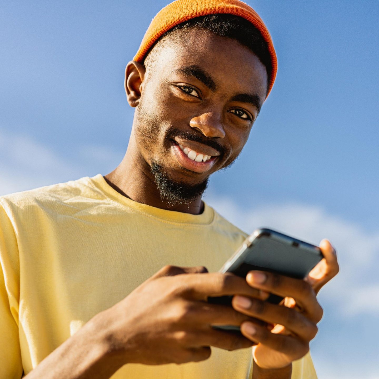 A smiling young man in a yellow t-shirt and orange beanie holding a smartphone against a blue sky.