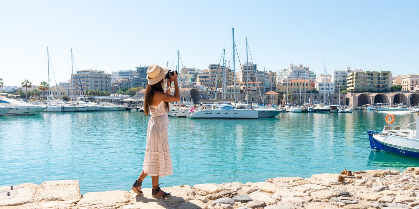 A woman standing at a harbor by crystal-clear blue water, taking a photo with her smartphone, with boats and the waterfront in the background on a sunny day. 