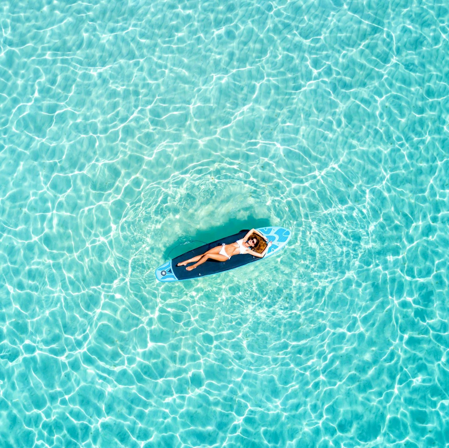 Woman in a bikini sunbathing on a surfboard in turquoise water.