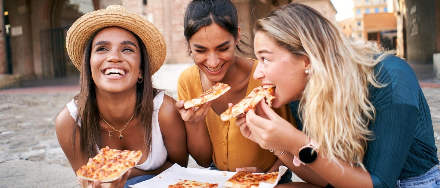 Funny mixed race group of three women eating street food outdoors in the city street. Happy female friends enjoying the summer vacations together European city. Young people lifestyle concept