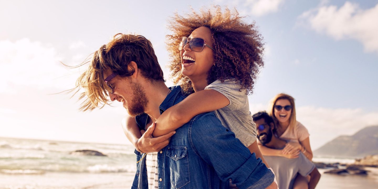 Group of friends walking along the beach, with men giving piggyback ride to girlfriends. Happy young friends enjoying a day at beach.