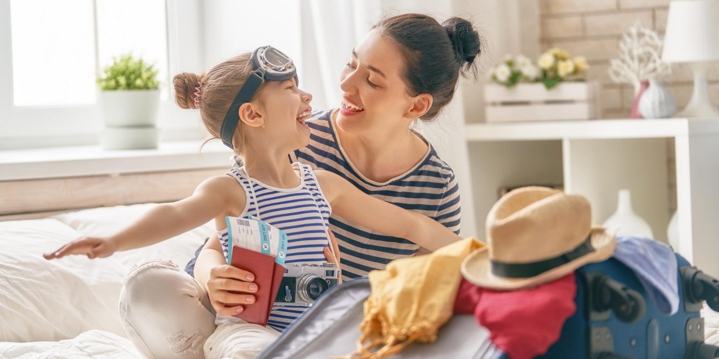 A joyful mother & daughter packing a suitcase for travel. The daughter, wearing goggles, holds a passport & camera.