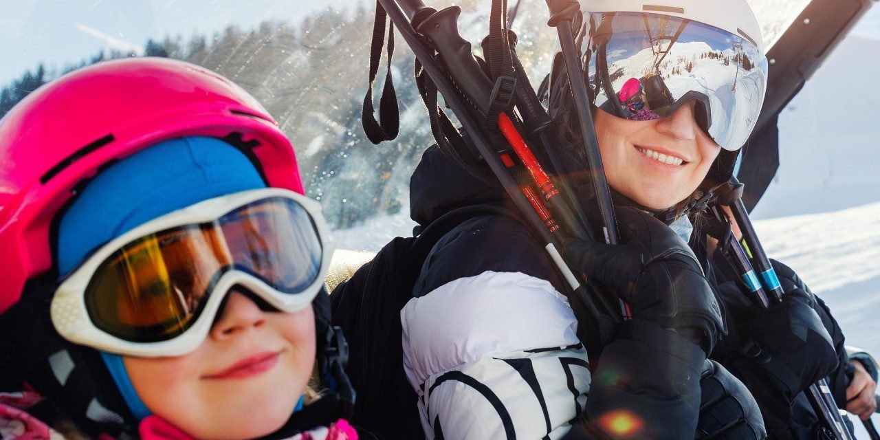 Two kids dressed in ski outfits, each holding a pair of skis, excited for skiing.