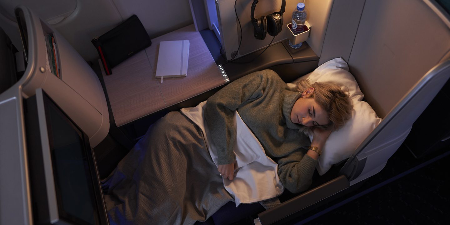 A person sleeps comfortably in a luxurious airplane seat, wrapped in a blanket. Nearby, a water bottle and headphones rest on a small table. Cozy ambiance.