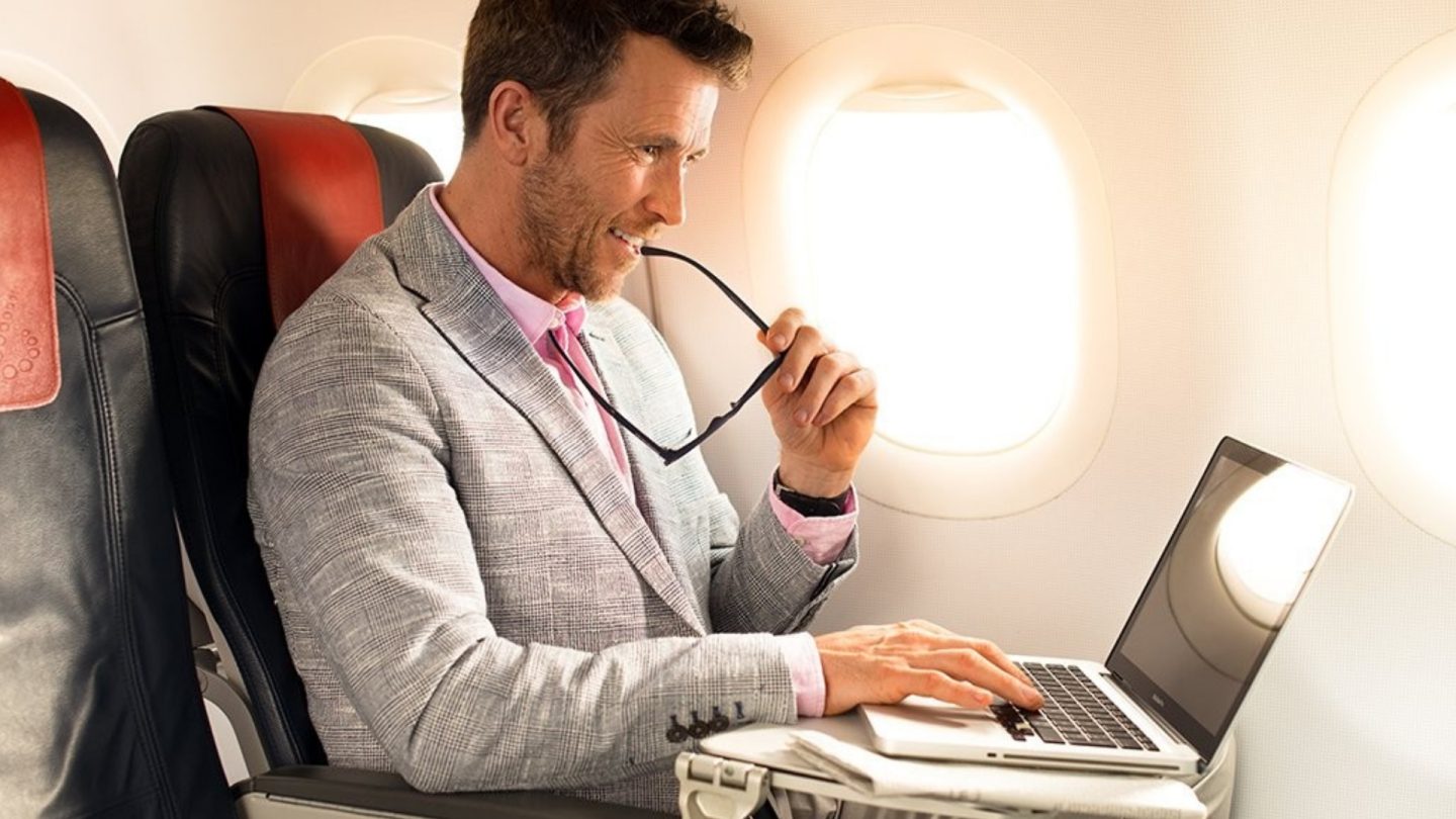 Smiling man working on laptop in an airplane window seat.