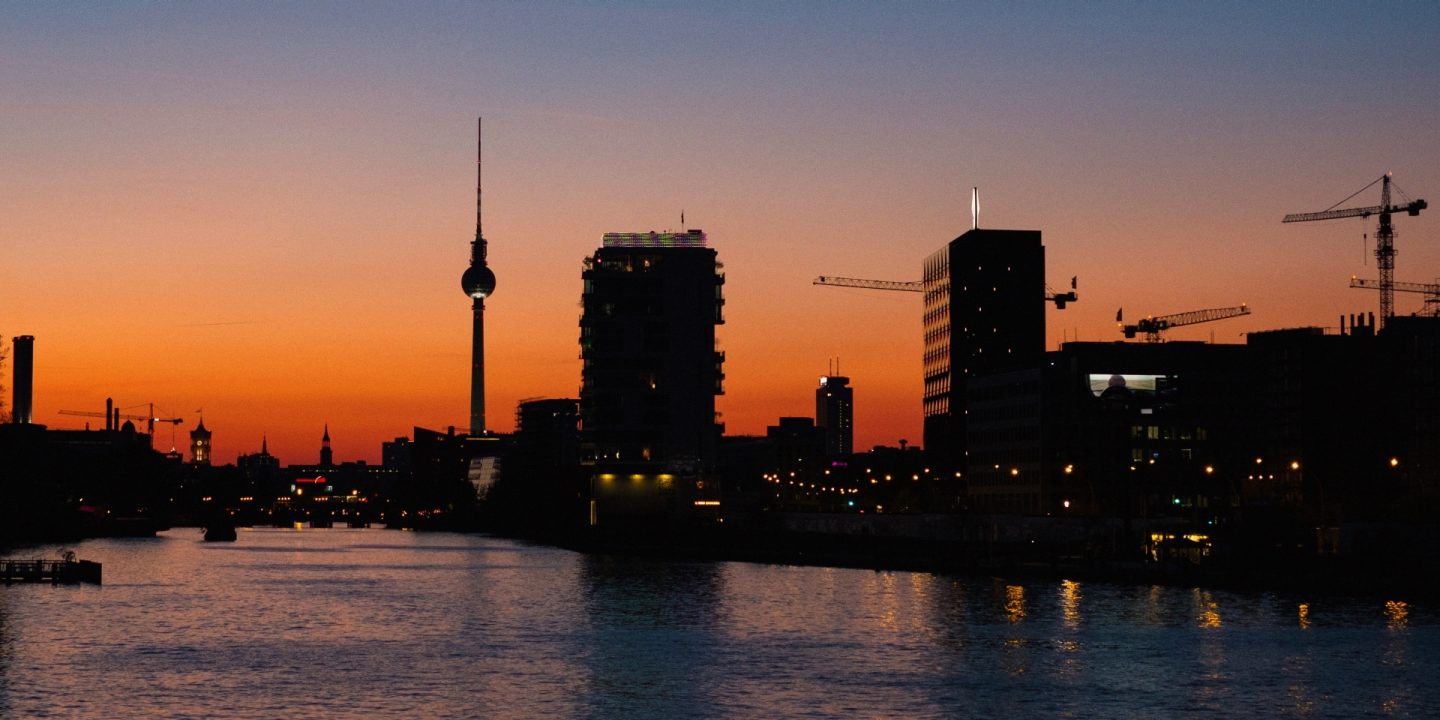 Stad bij zonsondergang met warme gouden en oranje tinten aan de hemel, gebouwen badend in zacht avondlicht.