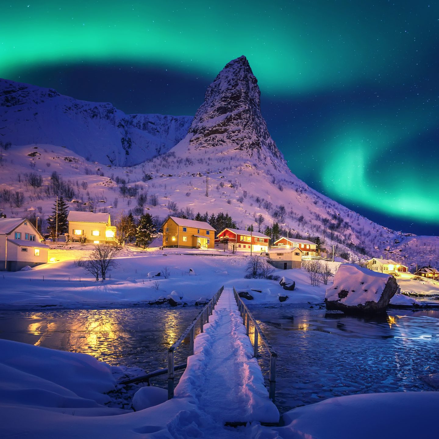 Des aurores boréales vertes au-dessus d’un village de montagne enneigé, avec des maisons colorées illuminées et un pont recouvert de neige enjambant une rivière gelée.