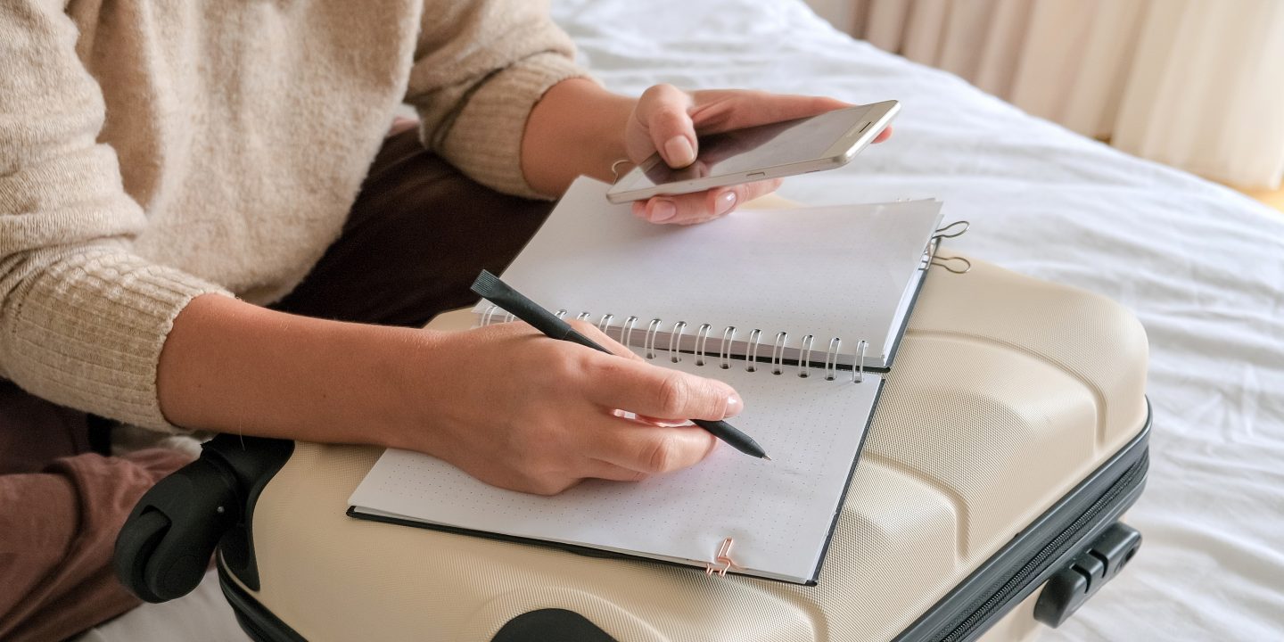 A traveler sits on a bed, planning a trip with a notebook and smartphone over a suitcase.