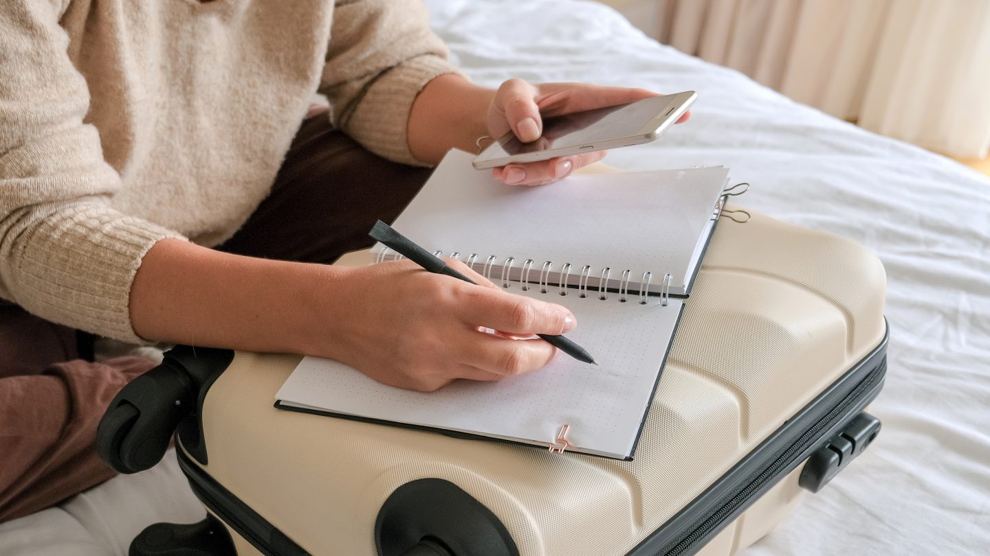 A person writing in a notebook on a suitcase while holding a phone.