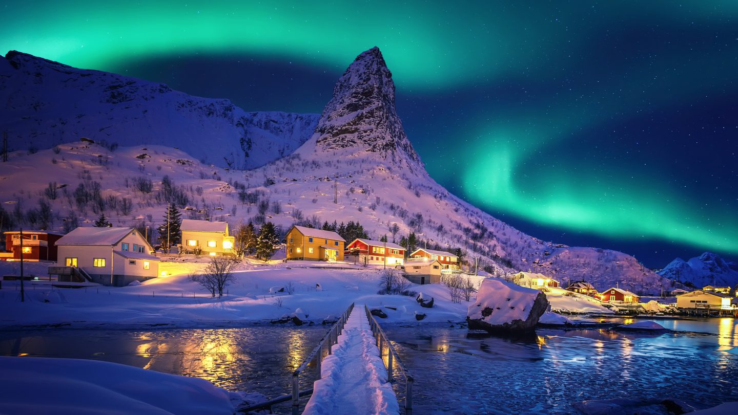 Green northern lights above a snowy mountain village with illuminated colorful houses and a snow-covered bridge over icy water.