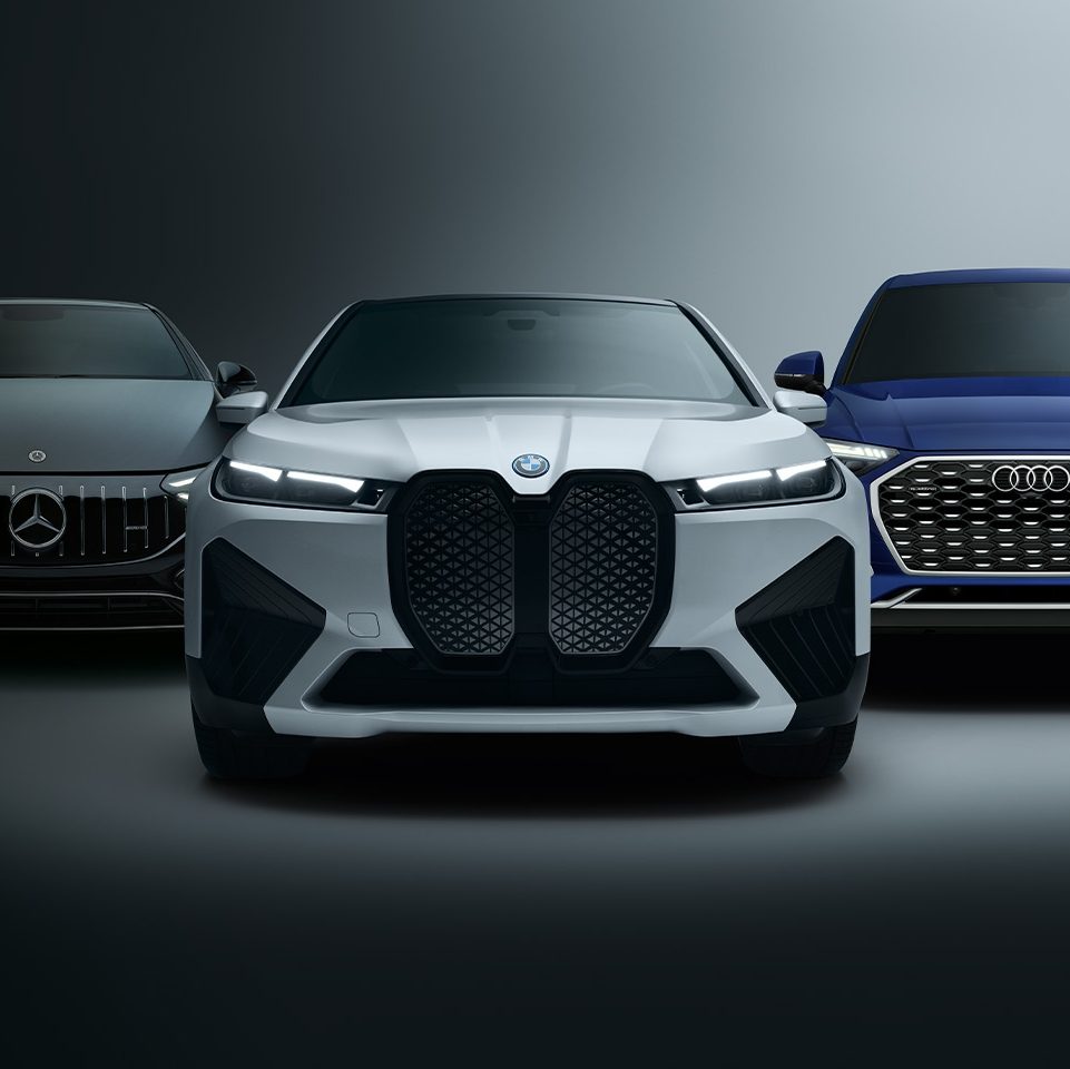 Front view of three cars from different brands against a dark background: a black Mercedes on the left, a white BMW in the center, and a blue Audi on the right.