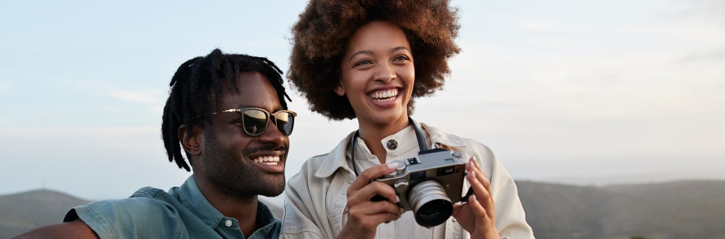 Happy young couple with camera in national wildlife park against sky during sunset